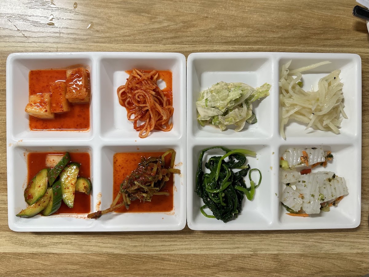 Eight side dishes laid out in two white divided trays, including kkakdugi radish kimchi, seasoned noodles, cabbage and cucumber